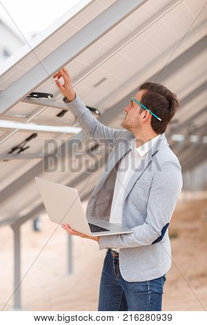 A man worker in a suit with a pencil behind his ear, holds a modern laptop in his hands and checks the solar panel. Outdoors