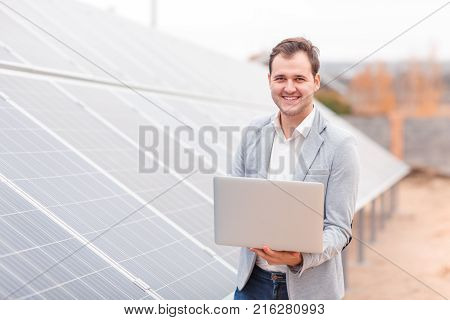 A smiling man, a solar cell employee, holds an open modern laptop in his hands standing next to the solar panel. Outdoors