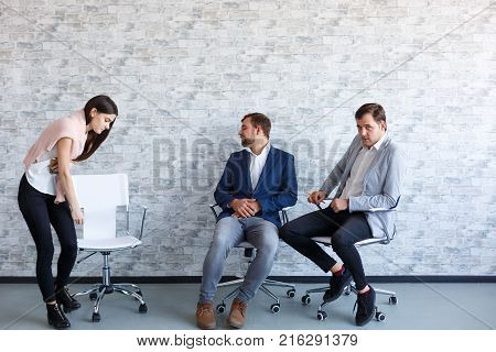 Two young men sit on chairs near a white stone wall, a young beautiful girl leans to an empty chair to sit down. Indoors.
