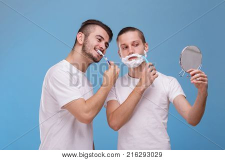Two young guys of European appearance look into one small round mirror. One of them brushes his teeth with a toothbrush, and the second shaves his beard with the help of shaving foam and a disposable razor. On a blue background.