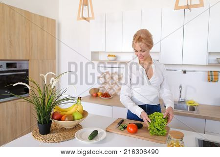 Attractive young woman cuts green cucumber for salad with sharp knife, takes red juicy tomato and several times throws up with smile on face, sniffs freshness of lettuce leaves and takes up shredding vegetables.  