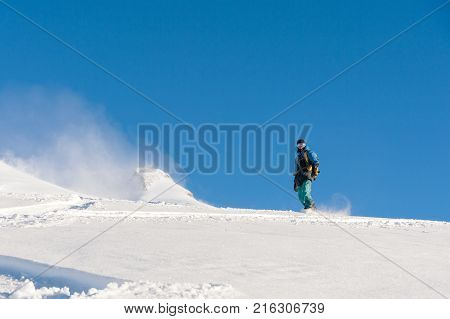 A freeboard snowboarder in a ski mask and a backpack skates over the snow-covered slope leaving behind a snow powder against the blue sky