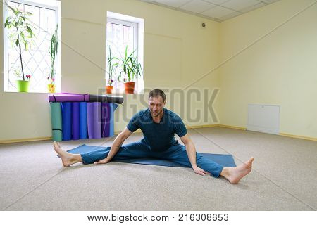 Young brunette of American origin sits on yoga mat and performs longitudinal twine. Short-haired sportswoman dressed in blue sports pants and blue t-shirt. Room light and spacious, under wall multi-colored yoga mats. Concept of to maintain body in good ph