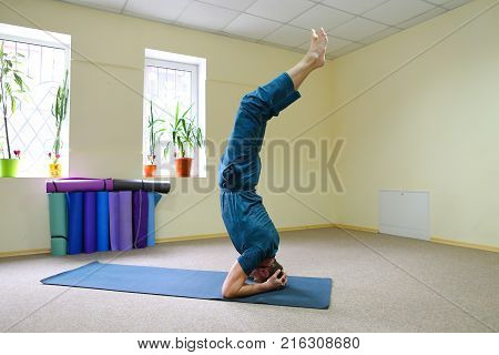 Young brunette of American origin sits on yoga mat and performs longitudinal twine. Short-haired sportswoman dressed in blue sports pants and blue t-shirt. Room light and spacious, under wall multi-colored yoga mats. Concept of to maintain body in good ph