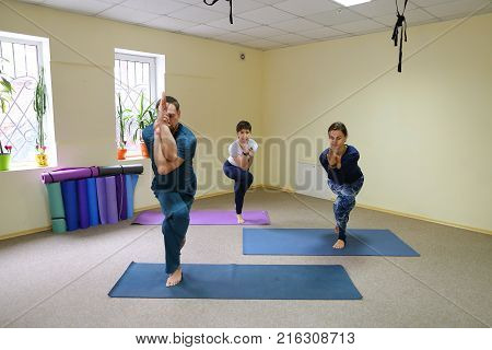 Two women and men of American appearance engaged in yoga, people sit on floor on fitness rugs and perform stretching exercises. Room light and spacious, walls of pleasant light color, on windowsills pots with flowers. Concept of maintain body in good phys