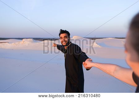 Young Muslim man leads sweet girl by hand, chatting with young woman and smiling, which goes for male. Lovers walk through expanses of sandy desert at sunset under open blue sky. Woman with fair hair tied in tail dressed in light t-shirt and blue jeans, d