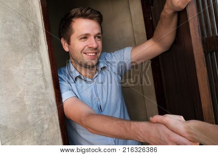 Portrait of young smiling man opening the door to his friend and welcoming him shaking hand. Invite somebody for party.