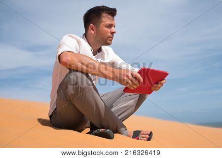 Lost in the desert. Caucasian man sitting on sand with tablet. He is lost and lookking in map where to go. Find way in fifficult situation