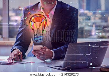 Businessman using smartphone and laptop at workplace with glowing polygonal lamp circuit supplies and other items. Blurry city view background. Innovation and creativity concept. Double exposure