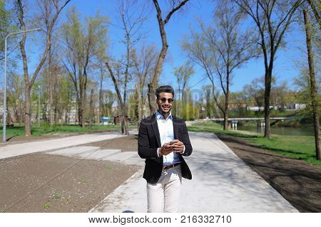 Guy walking in park and smiling, slow motion. Man dressed in blue shirt, white jeans and brown shoes. Boy has black hair and beard. Concept of places for rest and clean parks and cheeps fashion clothes for man