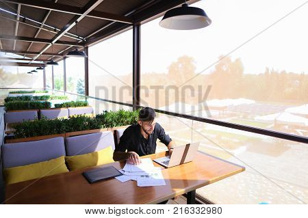 Freelance copywriter typing text fast on laptop keyboard at cafe table and looking tired. Handsome young man dressed in black shirt has dark hair and beard. Concept of typewriting skills and independent contractor for agencies.