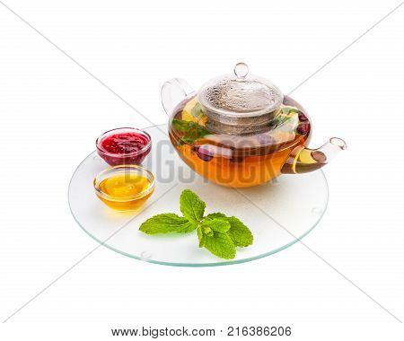 The glass teapot full of hot herbal tea with a dogrose costs on a glass opaque tray with saucers with honey and jam and mint leaves.
