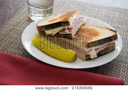A grilled ham and cheese sandwich on a white plate with a pickle beside glass of water and a napkin.