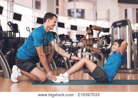 Father with his son in the same clothes in the gym. Father and son spend time together and lead a healthy lifestyle. Boy is pump the abdominals, coach helps him.