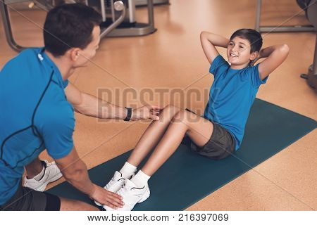 Father with his son in the same clothes in the gym. Father and son spend time together and lead a healthy lifestyle. Boy is pump the abdominals, coach helps him.
