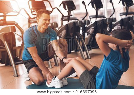 Father with his son in the same clothes in the gym. Father and son spend time together and lead a healthy lifestyle. Boy is pump the abdominals, coach helps him.