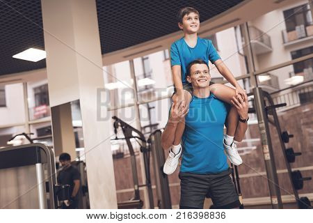 Father with his son in the same clothes in the gym. Father and son spend time together and lead a healthy lifestyle. The boy sits on the man's neck while he squats.