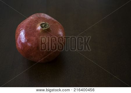 One large red pomegranate is placed on dark surface with natural light shining on it.