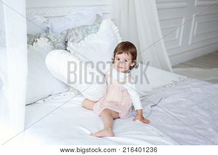 Funny and cute brunette little smiling girl playing on bed in bedroom. White interior with big bed