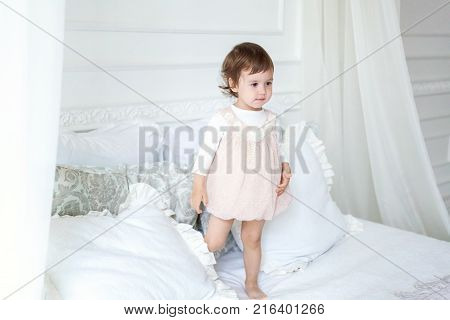 Funny and cute brunette little smiling girl playing on bed in bedroom. White interior with big bed