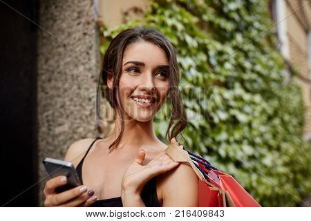 Close up portrait of young good-looking cheerful dark-haired caucasian girl looking aside with happy and relaxed expression, chatting with friends on phone, carrying shopping bags home after buying new clothes