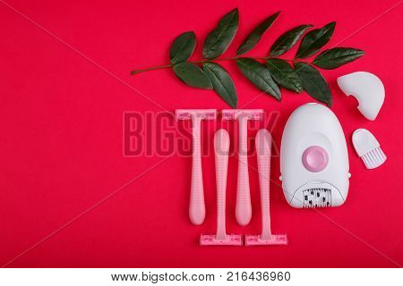 Close-up of a female epilator and disposable razor lie on a red background, next to it there is a green twig, a top view, a free place for text.