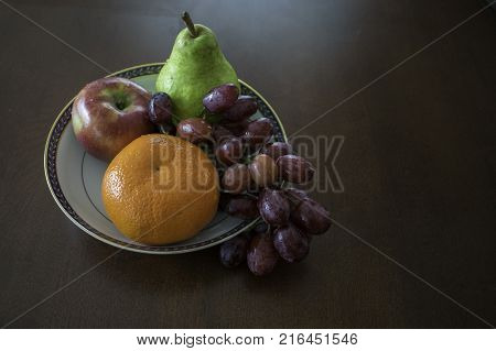 A selection of fruit consisting of a mandarin orange apple and grapes arranged in a white bowl on wood table top.