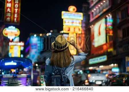 Young asian woman traveler with blue backpack and hipster hat looking night view on road with tuk tuk Thailand background at China Town Bangkok. Traveling in Bangkok Thailand