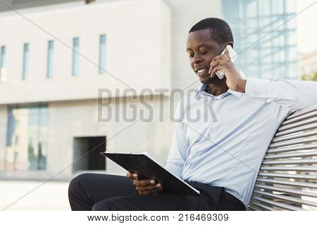 Serious black businessman talking on mobile and working with documents. African-american insuranse agent checking papers and consulting on smartphone, sitting on the bench outdoors in urban area