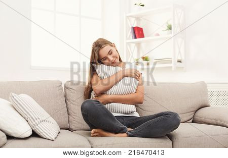 Happy dreamy girl sitting on beige couch and hugging her pillow. Thoughtful smiling young woman at home