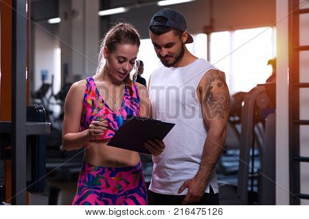 Happy smiling young woman standing with personal trainer in gym. Male coach and female client posing on the background of treadmills. Fitness and healthy lifesyle concept.