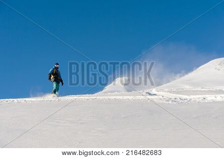 A freeboard snowboarder in a ski mask and a backpack skates over the snow-covered slope leaving behind a snow powder against the blue sky
