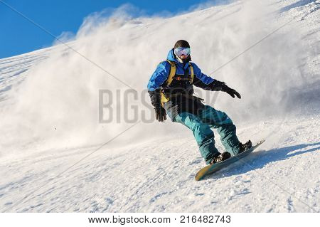 A freeboard snowboarder in a ski mask and a backpack skates over the snow-covered slope leaving behind a snow powder against the blue sky