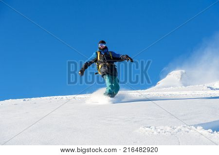 A freeboard snowboarder in a ski mask and a backpack skates over the snow-covered slope leaving behind a snow powder against the blue sky