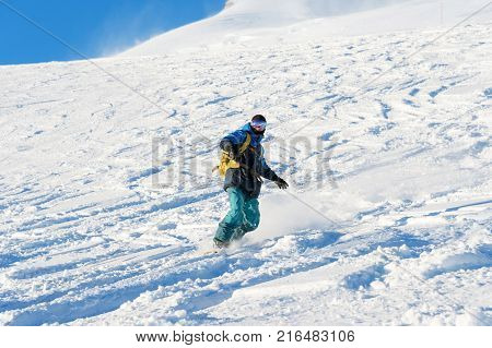 A freeboard snowboarder in a ski mask and a backpack skates over the snow-covered slope leaving behind a snow powder against the blue sky