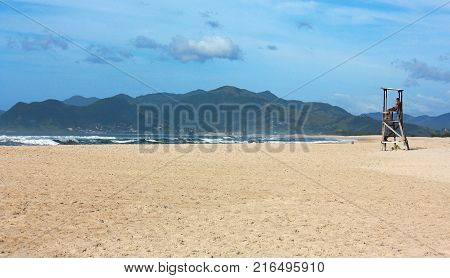 Lifeguard seated in his tower watching the sea, in the middle a deserted brazilian beach. The big beach in foreground, the sea and the mountains in background.