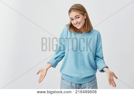 Indoor shot of beautiful Caucasian young female with straight fair hair shrugging her shoulders and smiling broadly feeling confused while refusing to go out on date with guy she doesn't like
