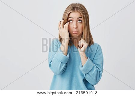 Portrait of caucasian young woman with oval face, dark eyes and fair straight hair wearing blue casual sweater, touching her face, trying to get eyelash out of her eye, looking upwards.