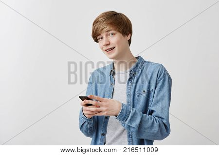 Trendy hipster male with fair hair and blue eyes wearing denim shirt posing indoors using cell phone, chatting with friends, typing messages. Clever student using modern technologies, looking at camera with smile