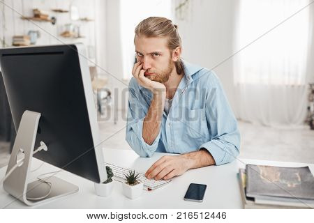 Bored exhausted young bearded Caucasian office worker with desperate look facing deadline but failing to finish report in time, looking with annoyance at camera. Male employee sitting in front of computer screen at light office, typing report.