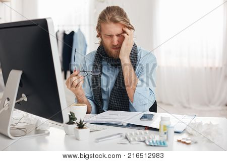 Sick and tired bearded male student or office worker has sleepy expression, runs temple because of feeling sick, surrounded by pills and drugs, tries to concentrate and finish work quicker