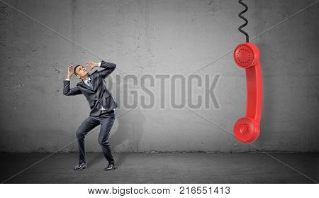 A small scared businessman on concrete background near a large red retro phone handle hanging down. Communications officer. Talking to clients. Negative feedback.