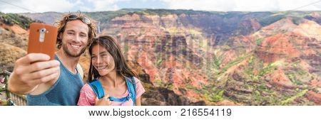 Hawaii couple taking selfie banner at Waimea Canyon Kauai. Hiking travel tourists taking mobile phone picture. Summer vacation people enjoying outdoor nature in Kauai, hawaiian islands.