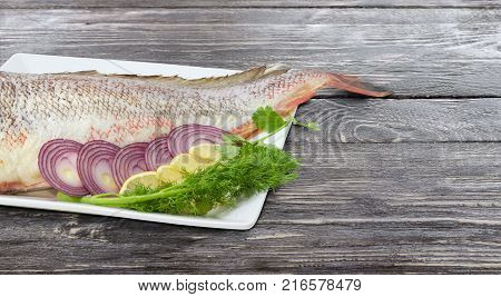 Fragment of the large square dish with uncooked red cod sliced lemon and red onion twigs of the dill and cilantro at shallow depth of field on an old rustic wooden table