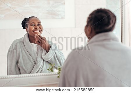 Smiling young African woman wearing a robe looking at her complexion in the mirror while standing in her bathroom in the morning
