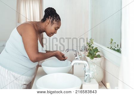 Young African woman washing her face wth water while standing over the basin in her bathroom in the morning