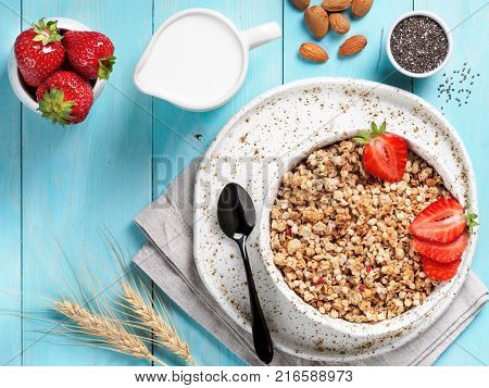 Top view of bowl with muesli, fresh strowberry, nuts, chia seeds and milk in milk jug on blue wooden background. Copy space.