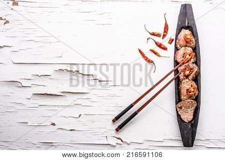 Slices of oriental spicy meat with sesame seads and pepper on long dark wooden plate with chopsticks the white wooden surface with cracked paint. Top view. Place for text.