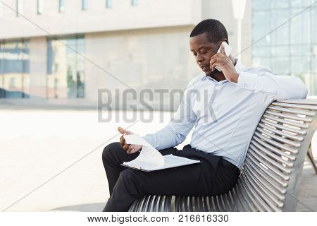 Serious black businessman talking on mobile and working with documents. African-american insuranse agent checking papers and consulting on smartphone, sitting on the bench outdoors in urban area