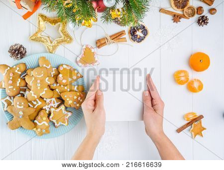 Top view female hands holding empty greeting card under white wooden table with gingerbred cookies, christmas tree branches with decor, spices. Selective focus. Text space
